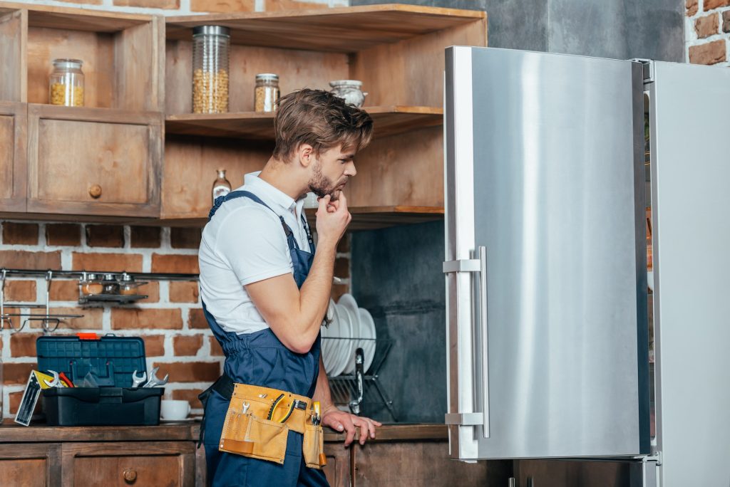 young repairman with tool belt looking at broken refrigerator